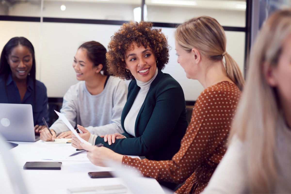 A group of people sit at a conference table while reviewing paper reports and laptop screens.
