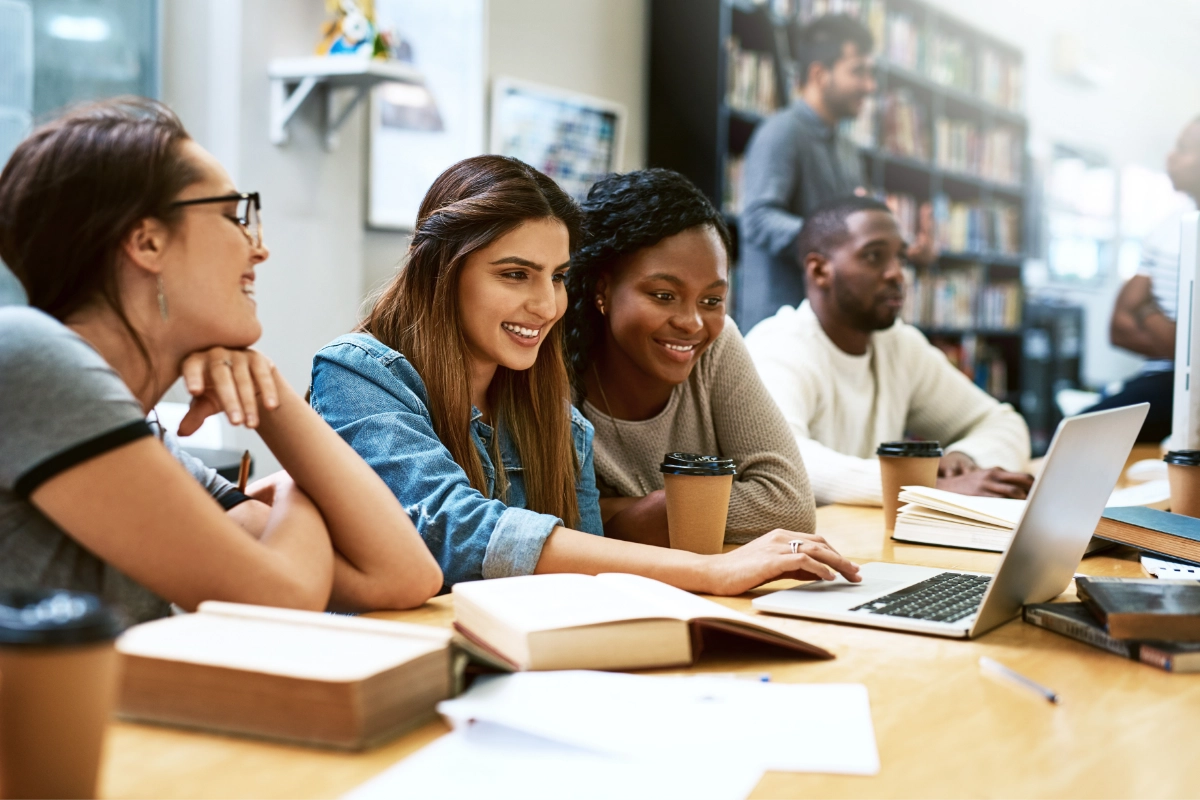 A diverse group of young adults sit at a table while looking at books, papers, and an open laptop.