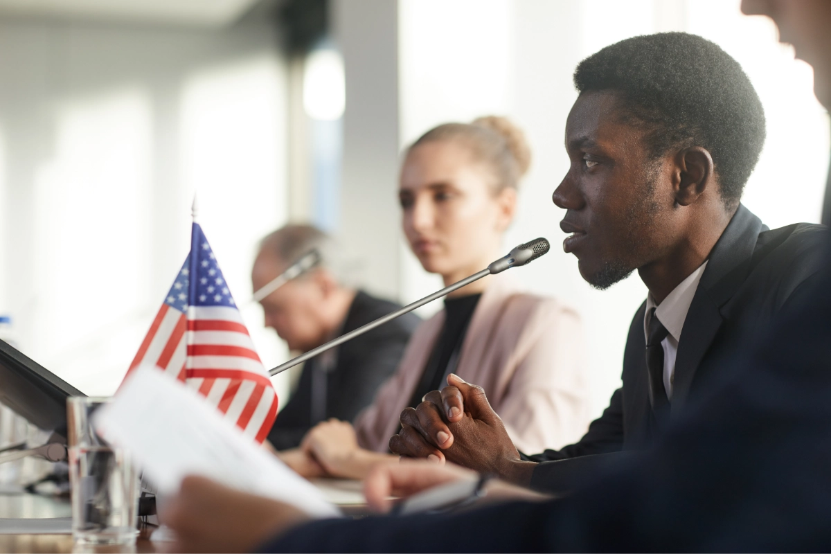 A person sits and speaks into a microphone at a conference or hearing beside an American flag and several other people.