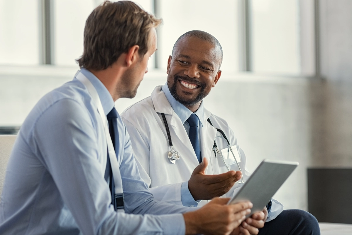 Inside a sunlit room, a person holding a notepad talks with another person wearing a white lab coat and stethoscope.