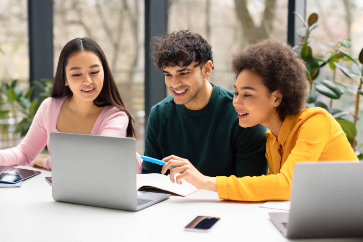Three young adults sit at a table, looking at a laptop together.