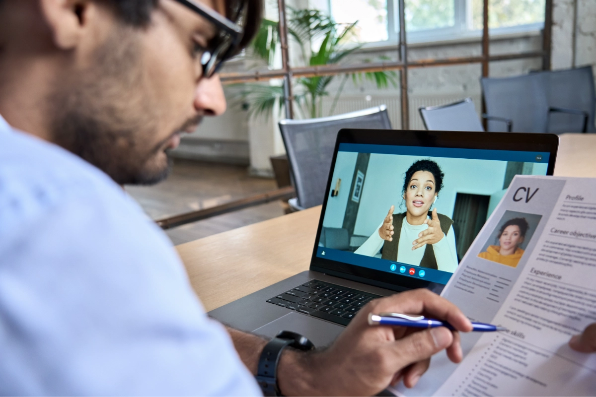 A team leader conducts an online interview with a potential hire using a laptop, while reviewing a printed copy of the applicant's CV.