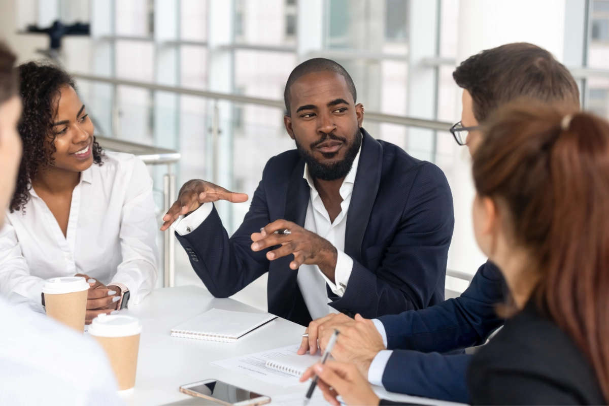 A diverse group of people sit at a conference table, take notes, and listen to a person at the center of the table speak.