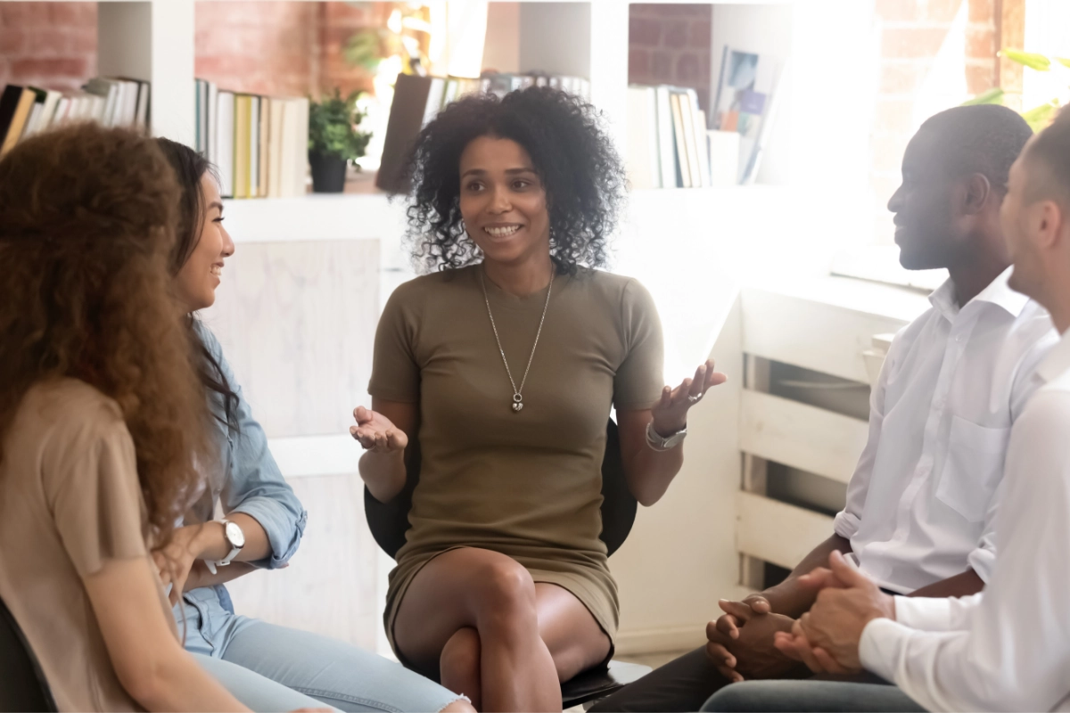 A small group of diverse individuals interact and sit on chairs arranged in a circle inside a sunlit office space.